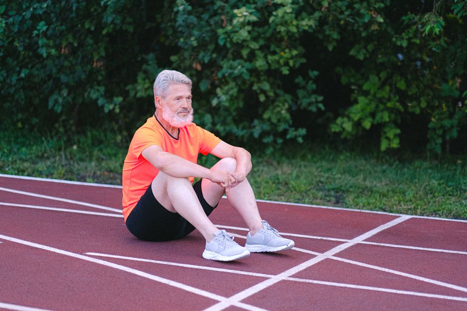 Side view of aged gray haired male in activewear looking away sitting on stadium track while having rest